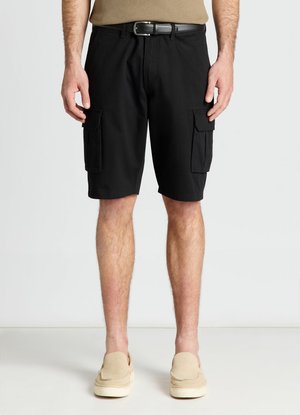 Uomo con pantaloncini cargo neri, scarpe beige, cintura nera e top beige, in piedi davanti a uno sfondo chiaro e uniforme.