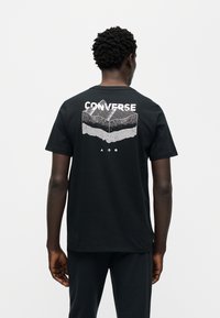 Μαύρο βαμβακερό μπλουζάκι με γραφικό βουνό σε λευκό στην πλάτη, με την επιγραφή "CONVERSE" πάνω σε έντονα γράμματα. Κοντά μανίκια, σχέδιο με στρογγυλή λαιμόκοψη.