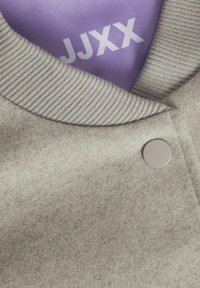 Ljusgrå, texturerad tyg med ribbad krage; har en silverknapp och lila innerfoder med vit text "JJXX."
