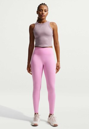 Kobieta stoi, ubrana w liliowy bezrękawnik typu crop top, różowe legginsy z wysokim stanem i białe buty sportowe na jednolitym tle.