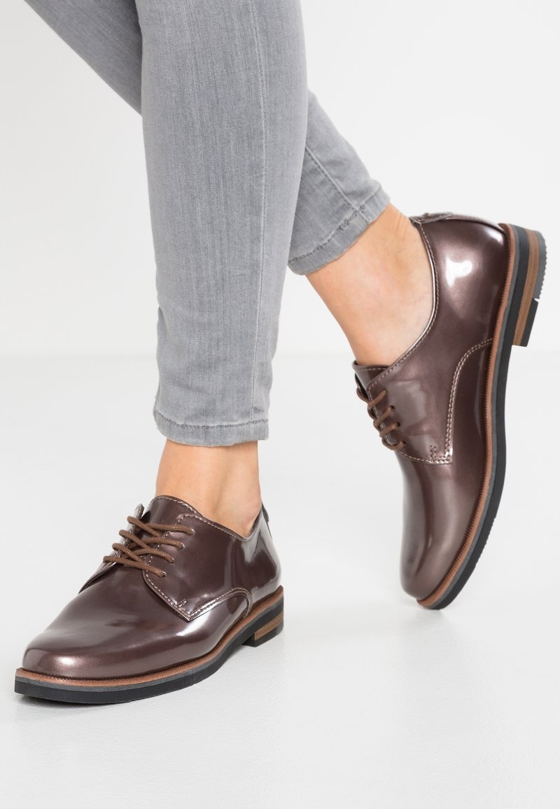 Chaussures en cuir marron à lacets avec une finition brillante, bout arrondi, et détail de semelle marron contrastante, portées avec un jean gris retroussé.