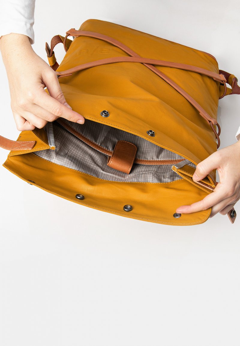 Gelbe Leder-Umhängetasche mit hellbraunen Lederträgern, ausgestattet mit einem karierten grauen Innenfutter und einem Druckknopfverschluss. Die Hand hält die Tasche teilweise geöffnet.