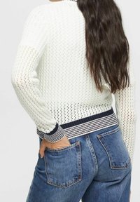 Weißer gestrickter Pullover mit strukturiertem Muster und einem marineblauen, gestreiften Rippbund. Kombiniert mit blauen Denim-Jeans.