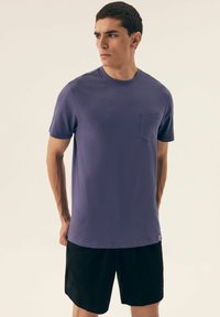 Fioletowa koszulka z krótkim rękawem, okrągłym dekoltem i kieszonką na piersi, zestawiona z czarnymi szortami. Tkanina wygląda na gładką i lekką.