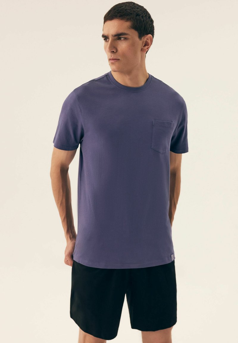 Fioletowa koszulka z krótkim rękawem, okrągłym dekoltem i kieszonką na piersi, zestawiona z czarnymi szortami. Tkanina wygląda na gładką i lekką.