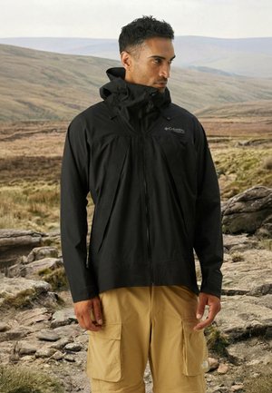 Mann trägt schwarze Columbia-Jacke und beige Cargohosen, steht auf felsigem Gelände mit sanft geschwungenen Hügeln im Hintergrund.