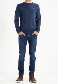 Pull à manches longues bleu marine avec col rond, associé à un jean bleu foncé. Bottines marron à lacets avec une semelle en caoutchouc. Détail discret du logo sur le pull.