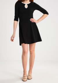 Abito nero con maniche a tre quarti, che presenta un dettaglio a chiave bianco nel collo e una gonna svasata. Modella che indossa scarpe beige con frange.