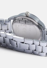 Sidabrinis nerūdijančio plieno laikrodis su grandinėlės apyranke, poliruotu paviršiumi, apvaliu korpusu ir ryškiu kronšteinu reguliavimui. Išgraviruota galinė dalis apima prekės ženklo detales.