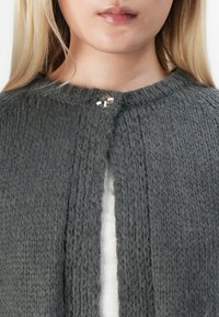 Blondýna v texturovaném tmavě šedém pleteném kardiganu zapnutém jedním stříbrným dekorativním knoflíkem přes bílý žebrovaný top.