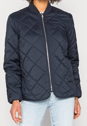 Femme portant une veste matelassée noire zippée avec un col côtelé et un jean bleu clair, debout devant un fond uni.
