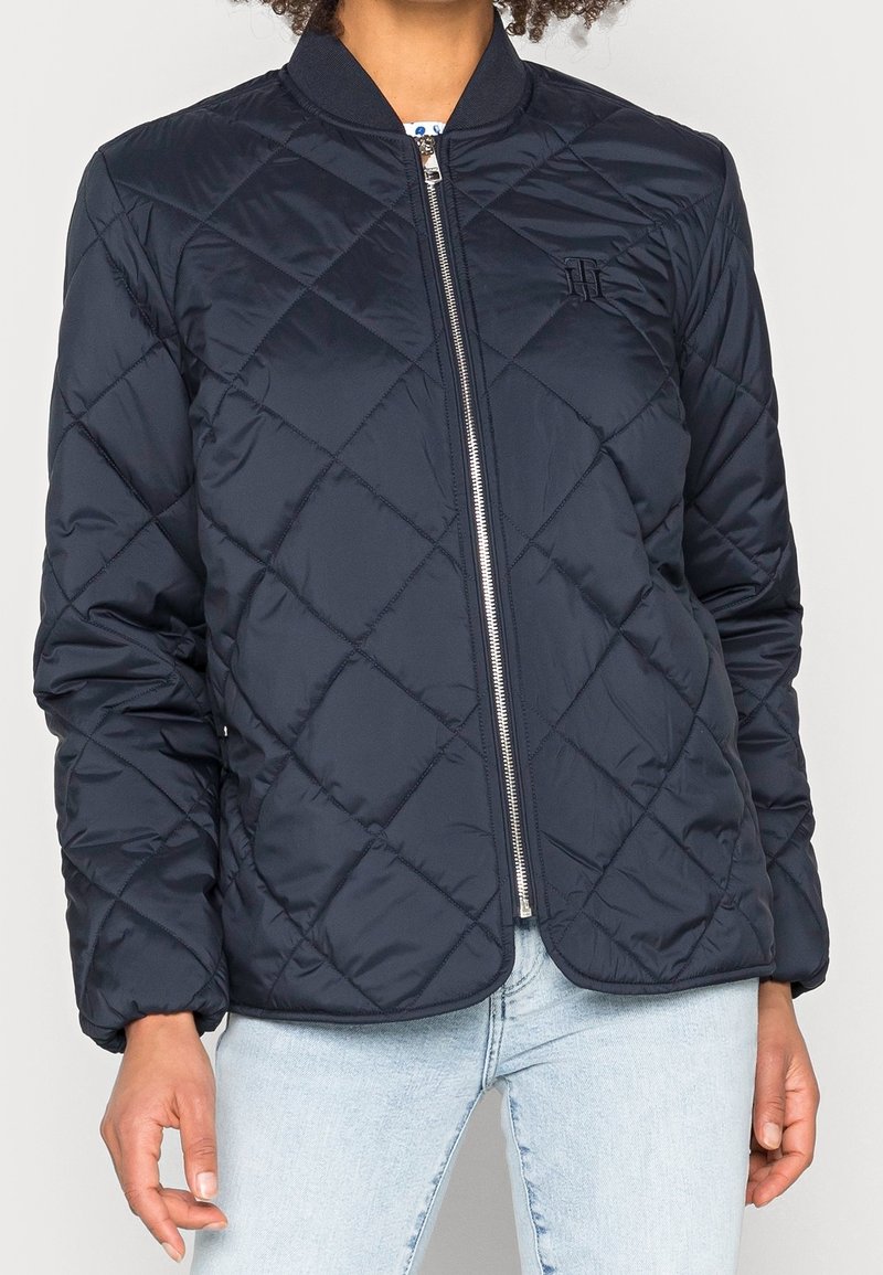 Femme portant une veste matelassée noire zippée avec un col côtelé et un jean bleu clair, debout devant un fond uni.