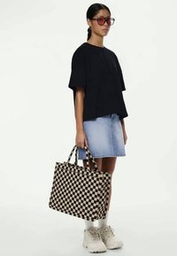 Črna in bež karirana torba z dvema zgornjima ročajema, jo nosi oseba v črnem zgornjem delu in jeans krilu. Bele robustne športne copate.