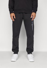 Sorte joggebukser laget av mykt stoff, med en avsmalnet design, elastisk midjebånd og en hvit logo langs venstre ben. Kombinert med hvite sko.