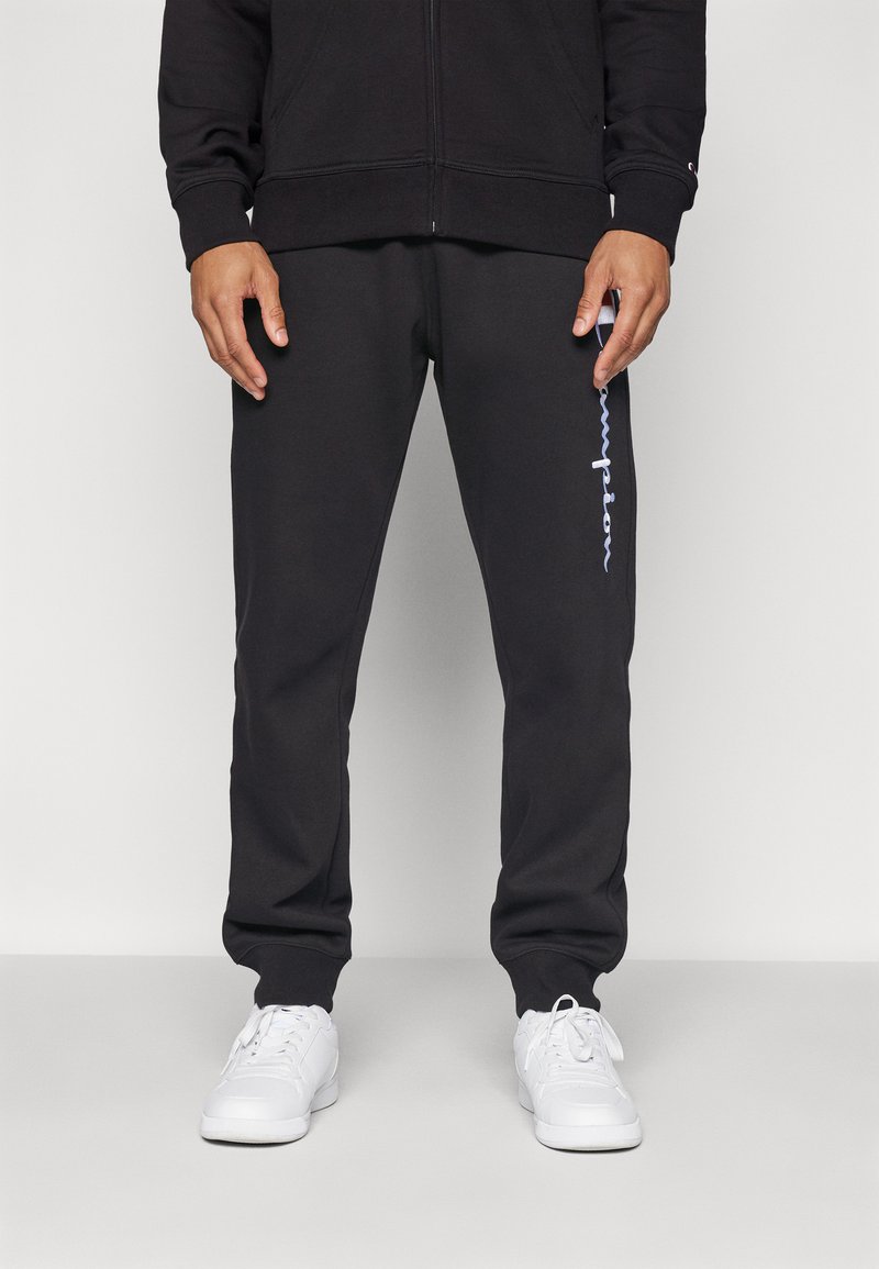 Sorte joggebukser laget av mykt stoff, med en avsmalnet design, elastisk midjebånd og en hvit logo langs venstre ben. Kombinert med hvite sko.