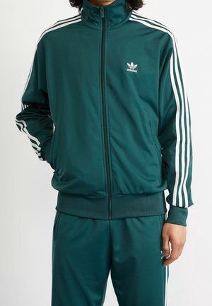 Person iført mørkegrønt Adidas træningssæt med hvide striber på ærmer og bukser, hænder i lommerne, lynlåsen foran lukket.