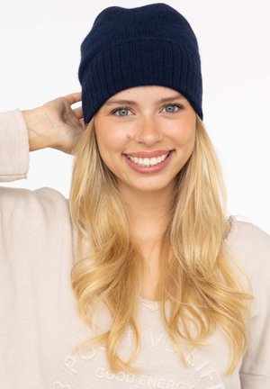 Navy knitted beanie with a ribbed texture, soft fabric, and rounded shape. Model wears a light sweater, showcasing natural hair and a smiling expression.