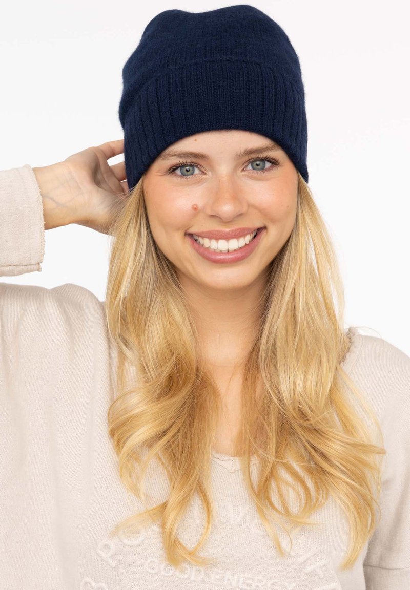 Navy knitted beanie with a ribbed texture, soft fabric, and rounded shape. Model wears a light sweater, showcasing natural hair and a smiling expression.