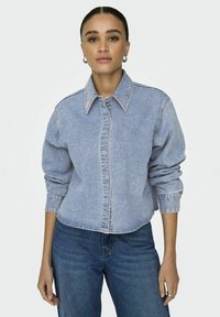 Light blue denim jacket with a collar, front buttons, and long sleeves. Features subtle speckles and a cropped hem. Worn over jeans.