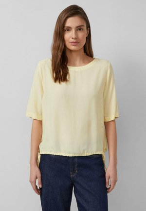 Kobieta o prostych brązowych włosach ubrana w jasnżółtą bluzkę z krótkim rękawem i ciemnoniebieskie dżinsy, stoi na tle jednolitego szarego tła.