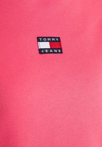 Růžové bavlněné tričko s černobílou obdélníkovou aplikací s nápisem "TOMMY JEANS" a barevným blokovým logem. Hladká textura, ležérní design.