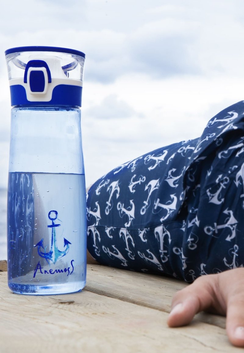 Klares Wasserflasche mit blauem Anker-Logo neben einer Person, die navyblaue Anker-musterisierte Hose trägt und die Hand auf einer Holzoberfläche im Freien ruhen lässt.