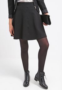 Czarna spódnica typu skaterek z teksturowanej tkaniny, noszona z czarnymi rajstopami i botkami za kostkę. Dopasowana talia, rozkloszowany krój i minimalistyczna kopertówka.