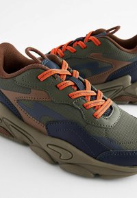 Grüne, braune und marineblaue Sneakers besitzen ein strukturiertes Gewebe und glatte Lederakzente, mit leuchtend orangefarbenen Schnürsenkeln und einer dicken Gummisohle.