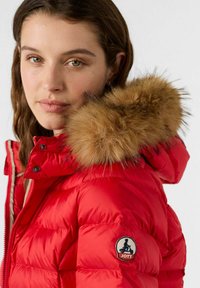 Giacca imbottita rossa con cappuccio bordato di pelliccia, tessuto trapuntato e toppa con logo sulla manica. Presenta chiusura a scatto e tasche laterali.