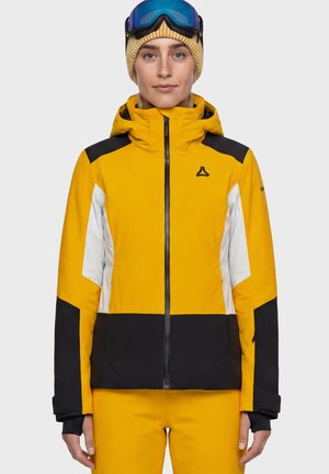 Person trägt eine gelbe, schwarze und weiße Skijacke mit Kapuze, passende gelbe Hose, beige Strickmütze und blaue Skibrille auf der Stirn.