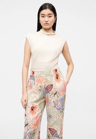 Γυναίκα με ίσια μαύρα μαλλιά που φοράει ασημένια σκουλαρίκια κρίκους, αμάνικο μπεζ τοπ και φαρδιά παντελόνια με φλοράλ μοτίβο, με τα χέρια στις τσέπες.