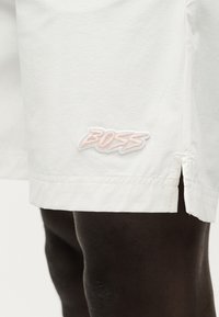 Λευκά σορτς με μικρή πλευρική σχισμή και ετικέτα με το λογότυπο "BOSS", φορεμένα από άτομο με σκουρόχρωμο δέρμα, ορατά από το μέσο του μηρού έως το γόνατο.