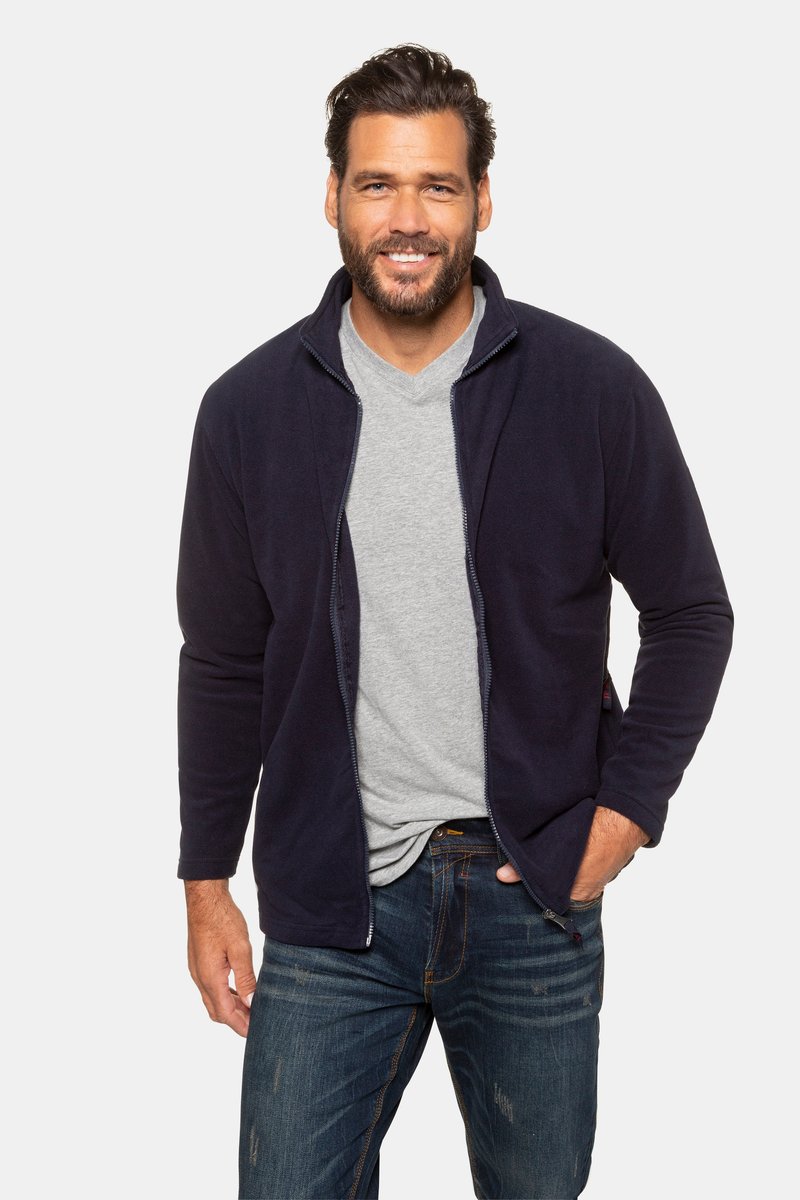 Mann mit dunklem Haar und Bart, der eine marineblaue Zipjacke, ein graues Shirt und blaue Jeans trägt, lächelt mit einer Hand in der Tasche.