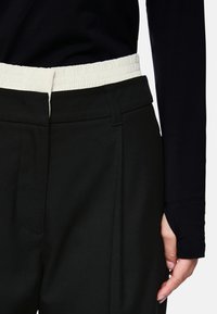 Person wearing black pants with a high-waisted white elastic waistband and a black long-sleeve top with thumb holes on the sleeve.