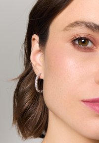 Przezroczyste kolczyki w kształcie koła z teksturowanym wzorem posiadają błyszczące akcenty. Kolczyki są noszone na uchu modelki, ukazując ich kształt i dopasowanie.