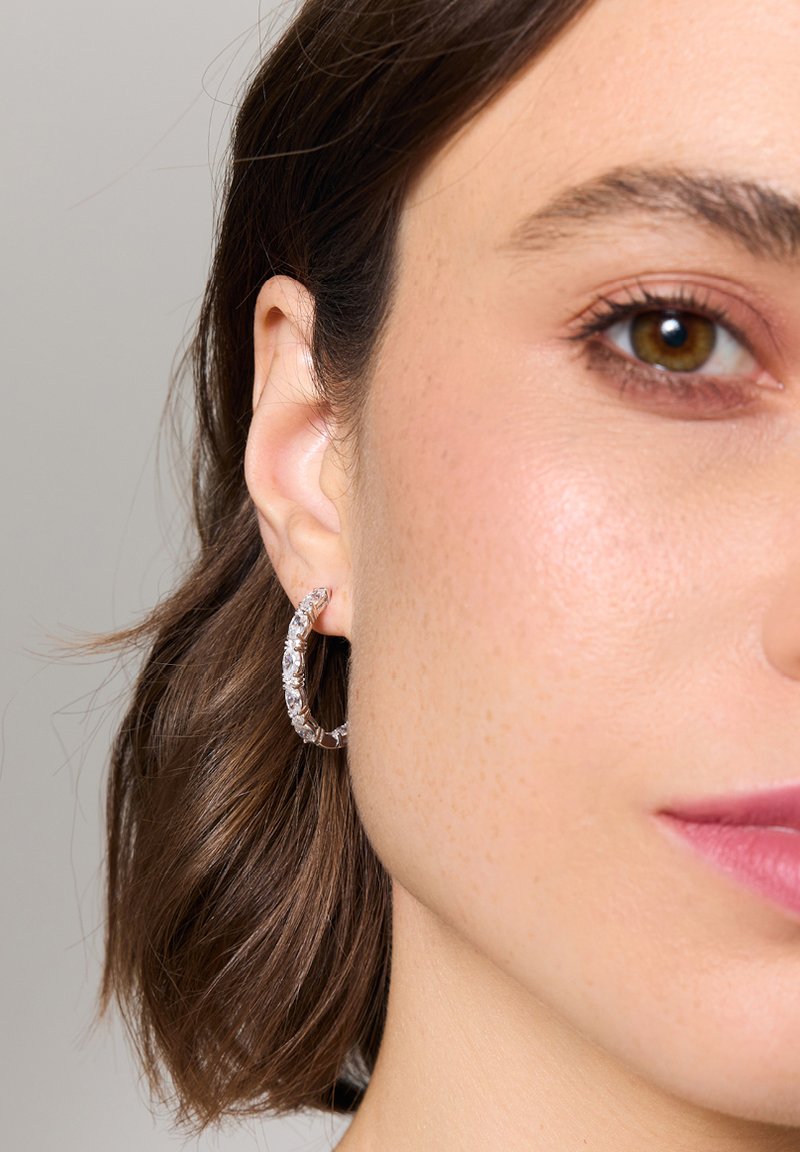 Przezroczyste kolczyki w kształcie koła z teksturowanym wzorem posiadają błyszczące akcenty. Kolczyki są noszone na uchu modelki, ukazując ich kształt i dopasowanie.