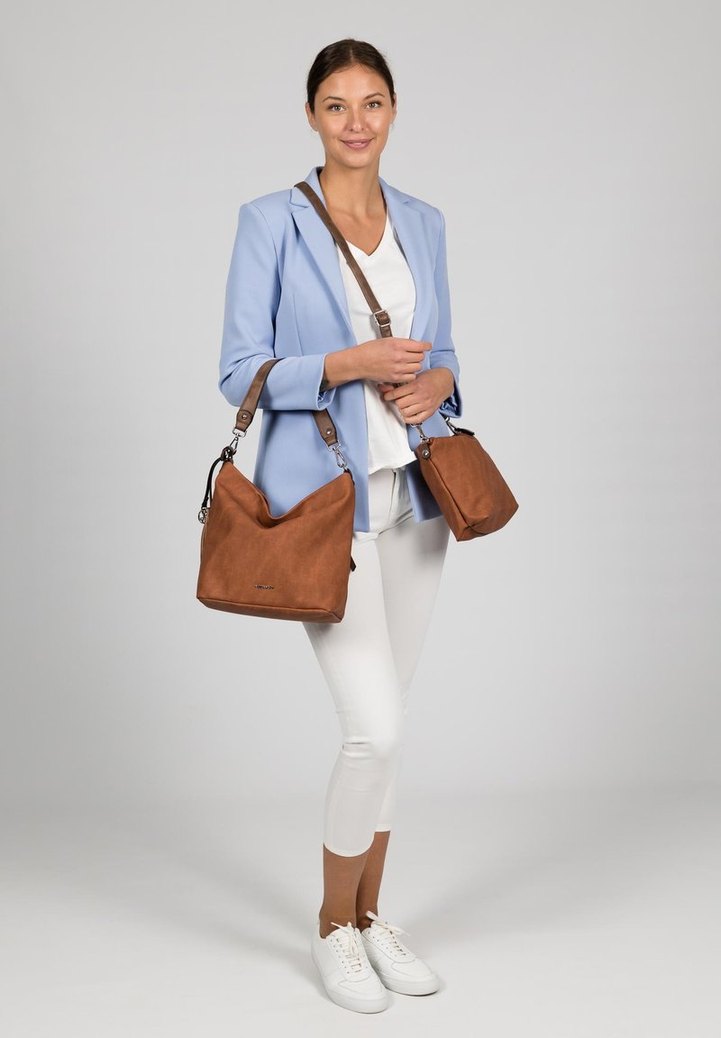 Sacs à épaule en cuir brun au design doux et décontracté, avec des accessoires en argent et des straps réglables, associés à un blazer bleu clair et un pantalon blanc.