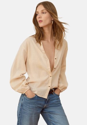 Vrouw met losse lichtbeige blouse en blauwe spijkerbroek, handen in de zakken, kijkt schuin met door de wind verwaaide haren tegen een effen achtergrond.