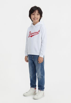 Usmievavý chlapec stojí oblečený v bielej mikine s nápisom "Dsquared²", modrých džínsoch a bielych vysokých teniskách na jednoduchom bielom pozadí.