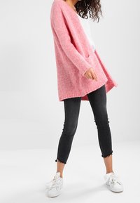 Pink knitted cardigan with long sleeves and open front, featuring two pockets. Worn over a white top and paired with black cropped jeans.