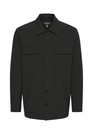 Person trägt eine schwarze Jacke mit Knöpfen, darunter einen dunkelblauen Reißverschluss-Pullover, blaue Jeans und schwarze Sneaker mit braunen Sohlen, steht mit einer Hand in der Tasche.