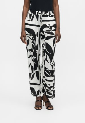 Osoba nosiaca čierno-biele nohavice s širokými nohavicami s abstraktnými lístkovými vzormi a čierne sandále na vysokom podpätku s remienkami.