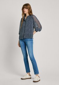 Marineblaue Bluse mit Blumenmuster, gerafften Ärmeln, Knopfleiste und feiner Textur, kombiniert mit hellblauen Slim-Fit-Jeans und weißen Sneakers mit braunen Akzenten.