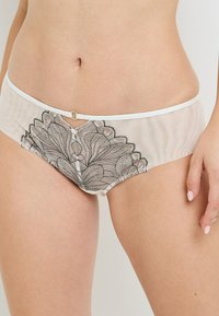 Panty hipster in rete bianca con dettagli intricati in pizzo floreale nero sul davanti, con un piccolo accento circolare al centro.