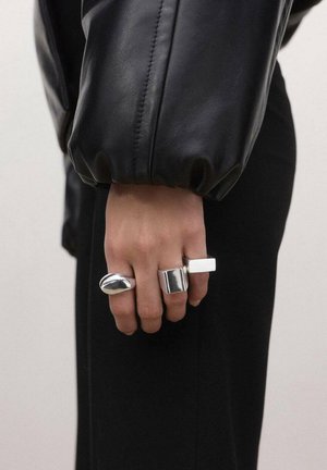 Roka s tremi debelimi srebrnimi prstani, oblečena v rokav črne usnjene jakne in črne hlače na svetli površini.