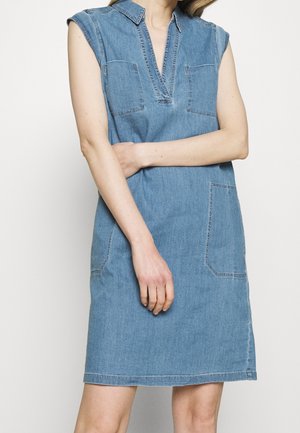 Kvinde iført ærmeløs blå denimkjole med krave, to brystlommer og en stor sidelomme, med armene krydset foran taljen.