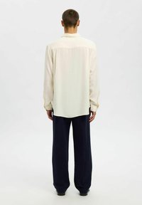 Camicia a maniche lunghe beige chiaro con colletto a punta, yoke posteriore e polsini abbottonati, abbinata a pantaloni navy larghi. Texture liscia.