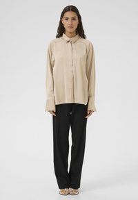 Beige satin skjorte med lange ærmer, krave, knaplukning foran og brede manchetter, sat sammen med sorte brede bukser og sandaler med stropper.