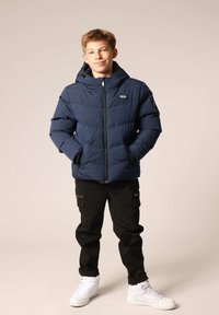 Marineblauwe pufferjack met een rits aan de voorkant, capuchon en zijzakken. Gecombineerd met zwarte cargobroek en witte sneakers. Glad, geïsoleerd materiaal.