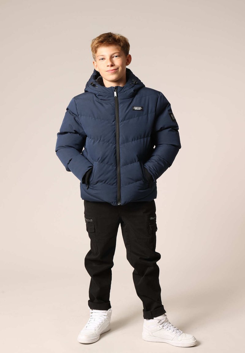 Marineblauwe pufferjack met een rits aan de voorkant, capuchon en zijzakken. Gecombineerd met zwarte cargobroek en witte sneakers. Glad, geïsoleerd materiaal.